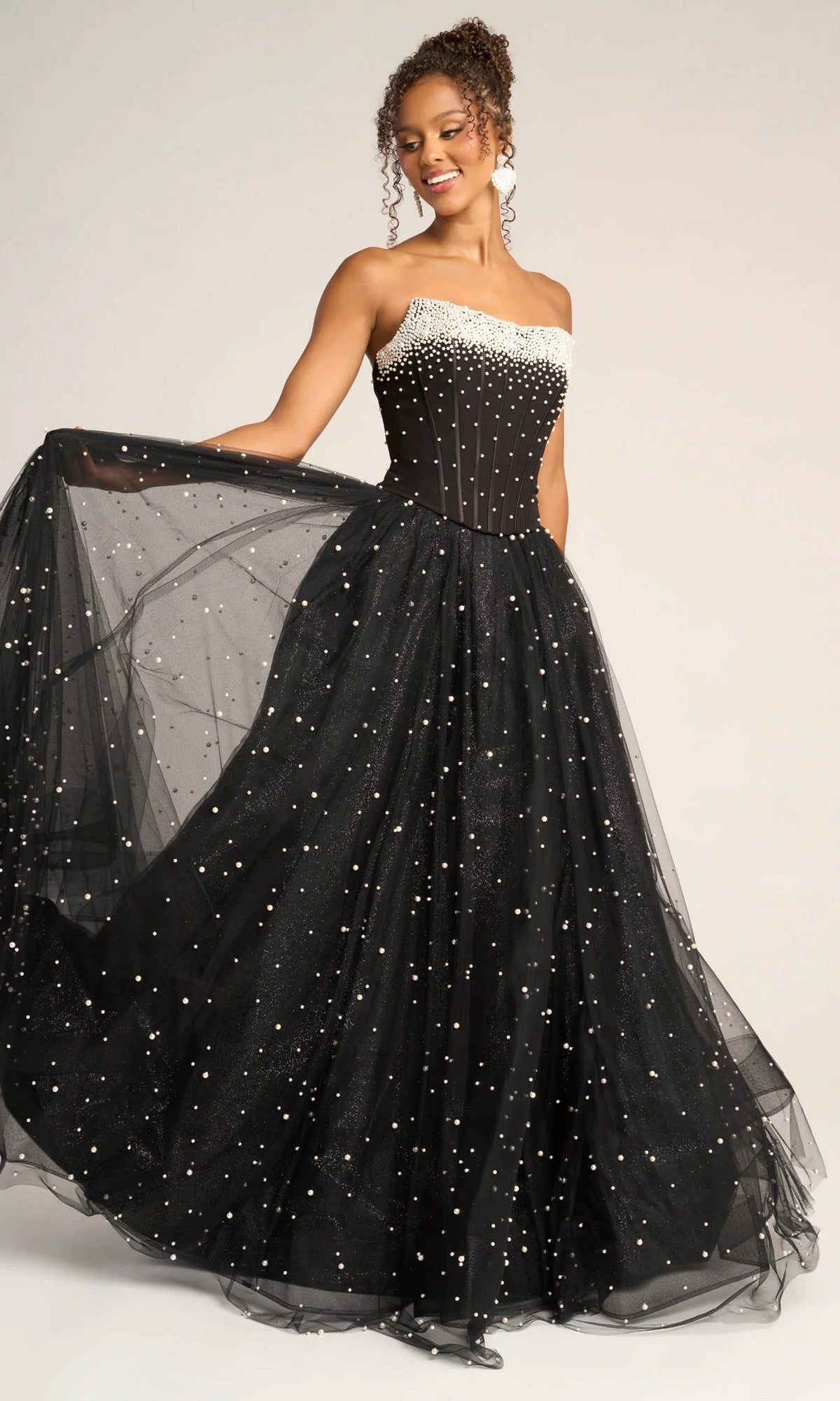 A woman wears the Portia and Scarlett PS26034 long formal dress in black, featuring a strapless corset bodice with pearl-like embellishments and a glitter tulle skirt, gracefully holding out one side.